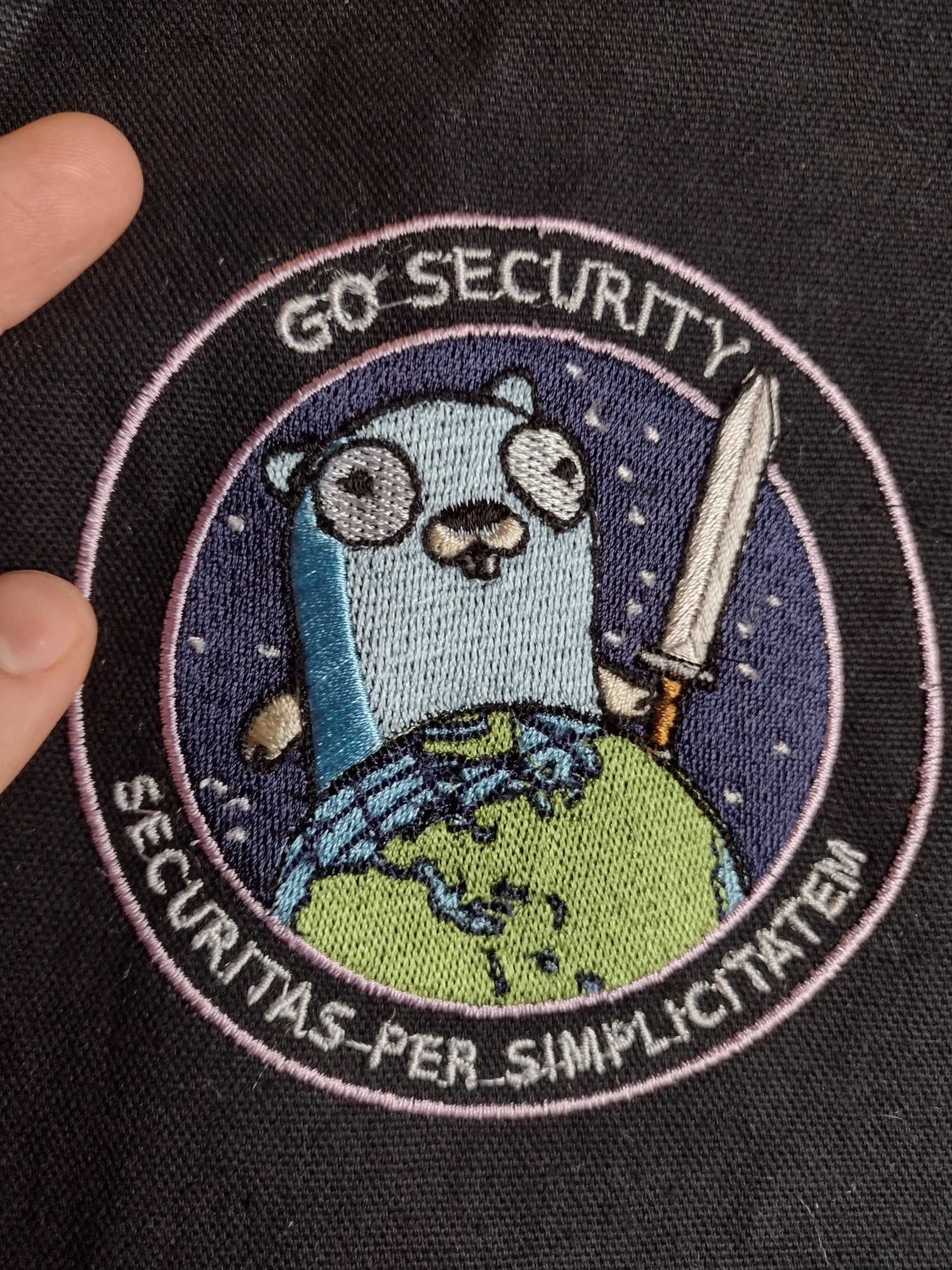 A machine-embroidered patch depicting a cartoon gopher holding a sword and towering over the planet earth. Around the border of the patch reads "Go Security" at the top and "Securitas Per Simplicitatem" at the bottom. The patch is four inches in diameter and inspired by the deranged National Reconnaissance Office NROL-39 mission patch.