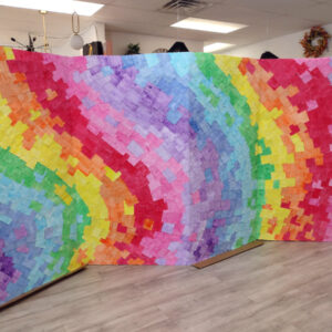 Photograph of a large 4-panel art piece consisting of almost a thousand pieces of rainbow-colored paper flowing across an unfolded refrigerator box. The work is set up in a brightly-lit shop.