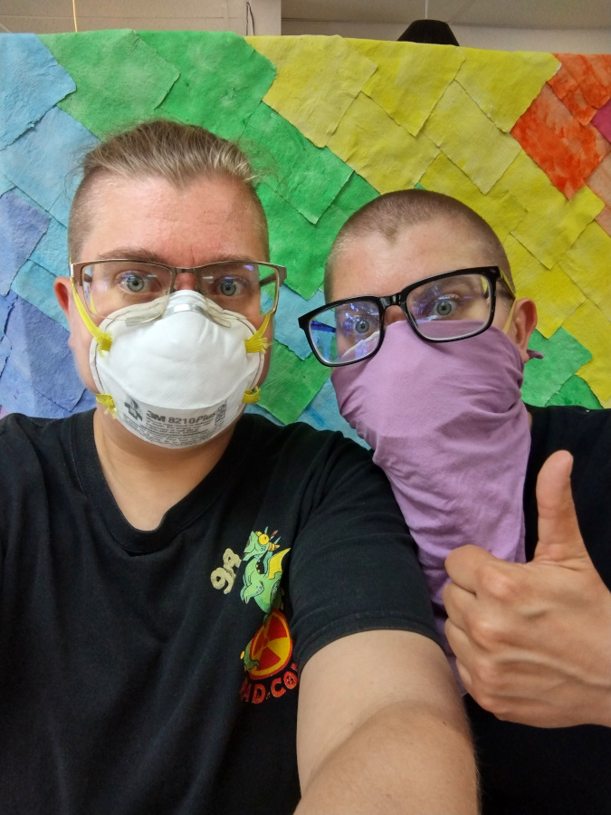 A photo of two suspiciously similar looking white people, both wearing masks and glasses, standing before the Rainbow Wall. L has long hair pulled back and wears an N95 mask, while Meg has a buzz cut and wears a purple PFLAG bandana over their mask. L holds up their phone for the photo while Meg gives a thumbs up.