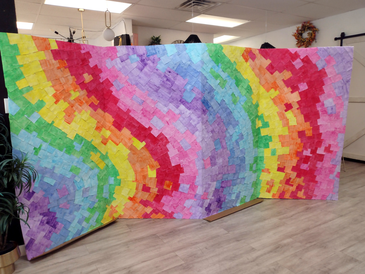 Photograph of a large 4-panel art piece consisting of almost a thousand pieces of rainbow-colored paper flowing across an unfolded refrigerator box. The work is set up in a brightly-lit shop.